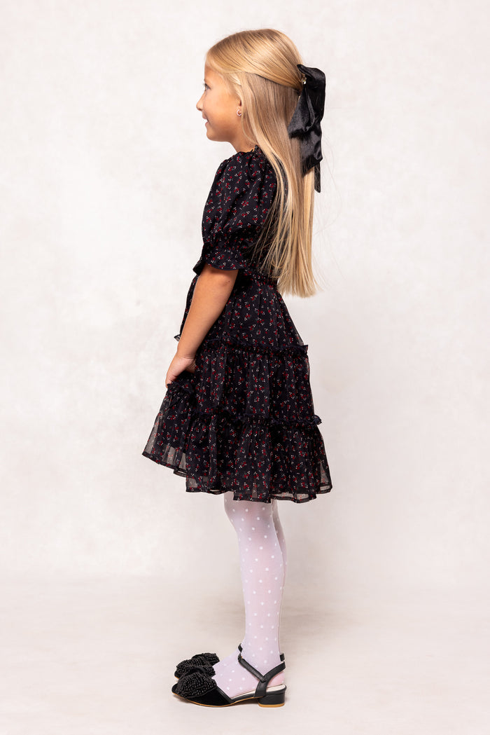 Young girl wearing a black floral dress with white tights and black shoes on a plain background