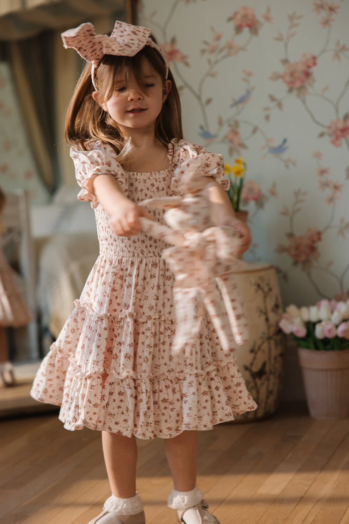 A young girl in the Ivy City Co Mini Madeline Dress in Pink and a matching bow headband twirls with a soft toy in a sunlit room, floral wallpaper and tulips brightening the cozy background.