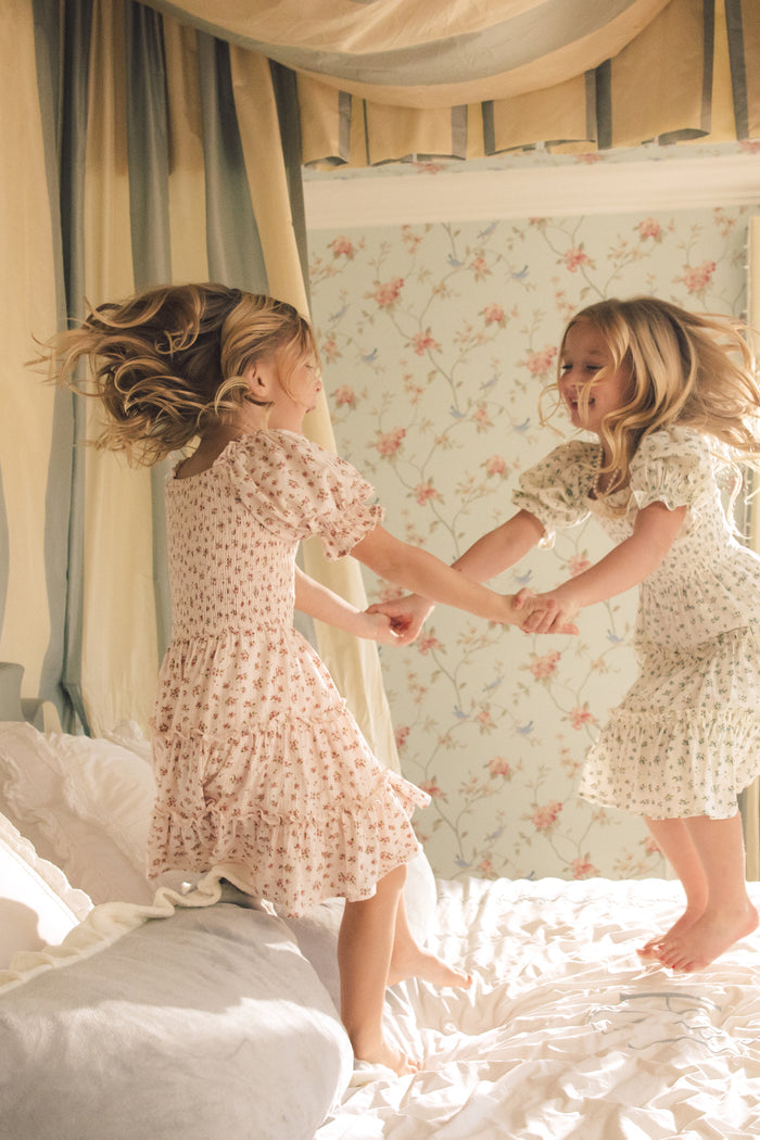 Two young girls joyfully hold hands and jump on a bed, each wearing the Ivy City Co Mini Madeline Dress in Pink, in a softly lit room with floral wallpaper and a canopy over the bed.