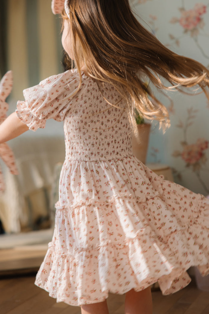 A young girl with long brown hair twirls joyfully indoors in the Ivy City Co Mini Madeline Dress In Pink, a floral cotton dress with ruffles. Her hair and dress flow outward as she spins in a softly lit room with floral wallpaper.