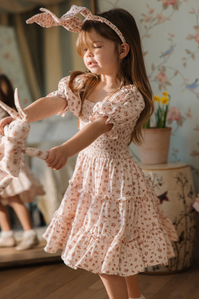 A young girl in an Ivy City Co Mini Madeline Dress in Pink, a light pink floral cotton dress with matching headband, twirls indoors with a fabric bunny amid natural light, floral decor, a mirror, and potted flowers.