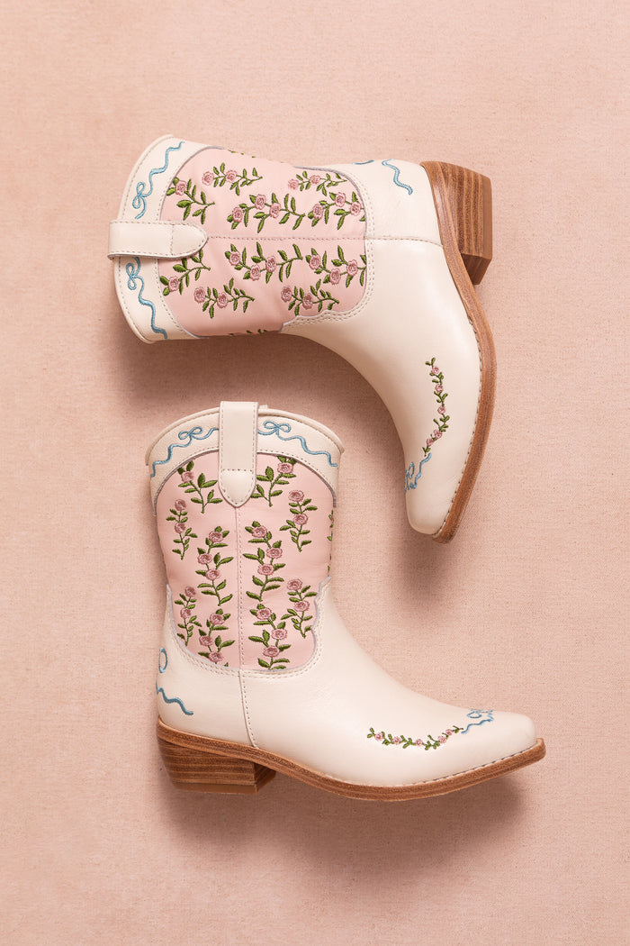Two Mini Rosemary Cowgirl Boots in Cream & Pink by Ivy City Co, featuring wooden heels, floral embroidery, and delicate pink trim, are placed on a soft pink background.