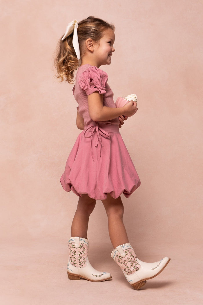 A young girl stands sideways, smiling in Ivy City Co’s Mini Rosemary Cowgirl Boots in Cream & Pink, paired with a matching pink dress and holding a coordinating doll. She wears a white bow in her hair, set against a soft pink background.