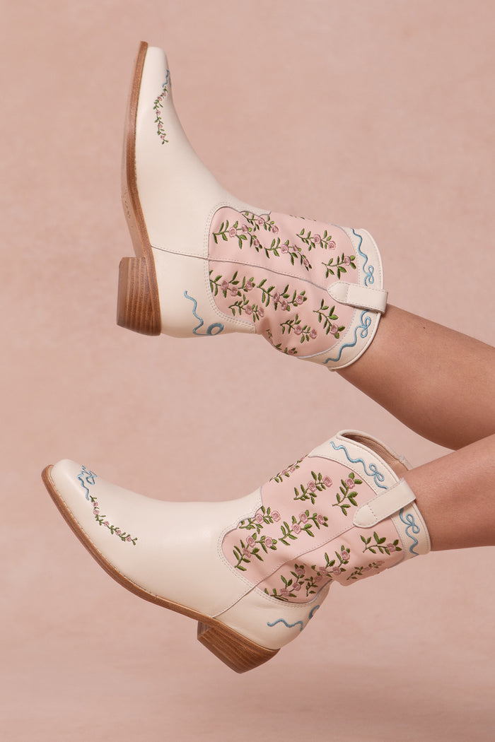 A pair of cream and pink cowgirl boots with floral embroidery, pink trim, and wooden heels, modeled by a person lying down with legs raised against a soft pink background.