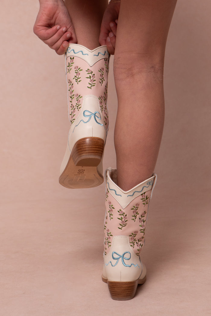 A person stands on tiptoe against a light background wearing Ivy City Co’s Mini Rosemary Cowgirl Boots in Cream & Pink, featuring floral embroidery and light blue bows.