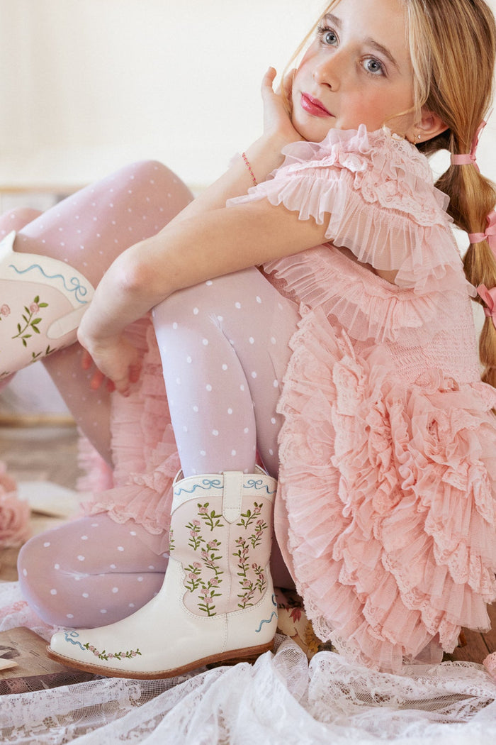 A young girl in a pink ruffled dress and polka dot tights sits with her knees up, wearing Ivy City Co’s Mini Rosemary Cowgirl Boots in Cream & Pink. Her blonde hair is styled in a braid, and she gazes thoughtfully to the side.