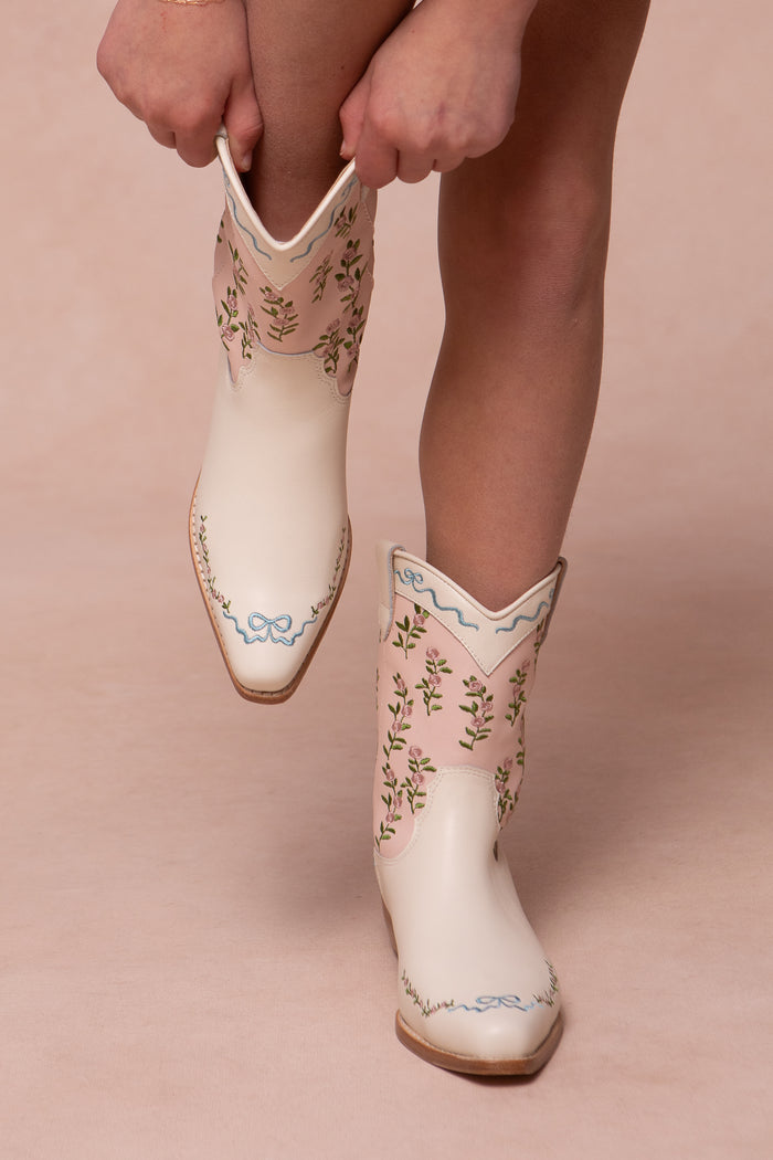 Wearing the Ivy City Co Mini Rosemary Cowgirl Boots in Cream & Pink—featuring floral embroidery, small blue bows, and delicate pink trim—a person stands on a light pink background.