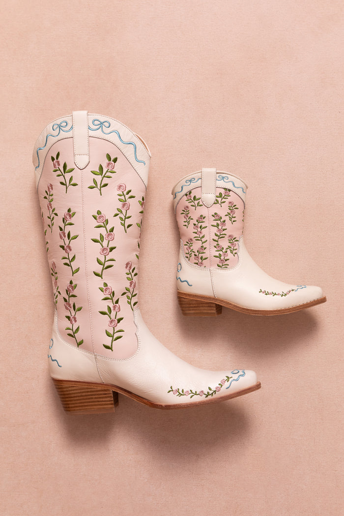 Two cream-colored Mini Rosemary Cowgirl Boots by Ivy City Co, featuring pink and green floral embroidery and blue ribbon accents on wooden heels, displayed on a pale pink background.