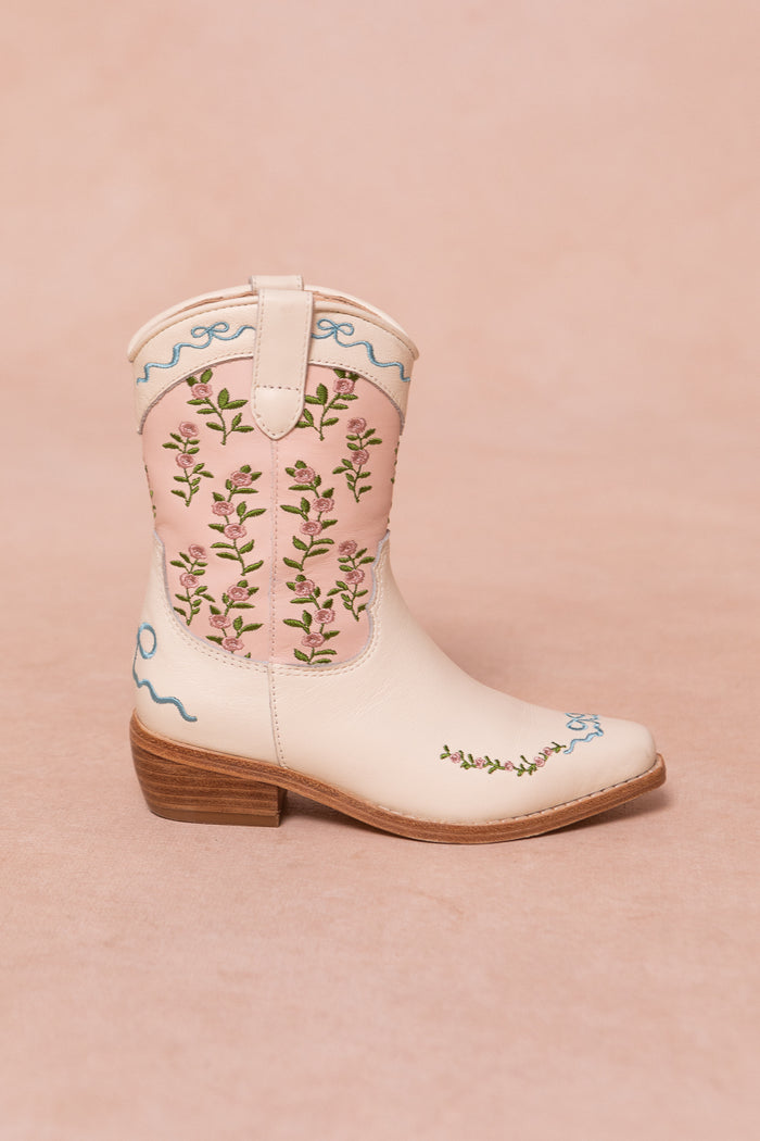 Introducing the Mini Rosemary Cowgirl Boots in Cream & Pink by Ivy City Co: these cream-colored children’s boots feature a light pink panel adorned with embroidered pink flowers and green leaves, accented by blue decorative stitching and playful pink trim. The classic white leather-inspired design is finished with a wooden heel—all perfectly showcased against a soft pink background.