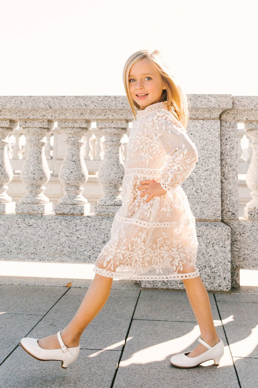 A young girl with blonde hair confidently walks outdoors in sunlight beside a stone balustrade, wearing the Ivy City Co Mini Sicily Dress with Long Sleeves and white shoes.