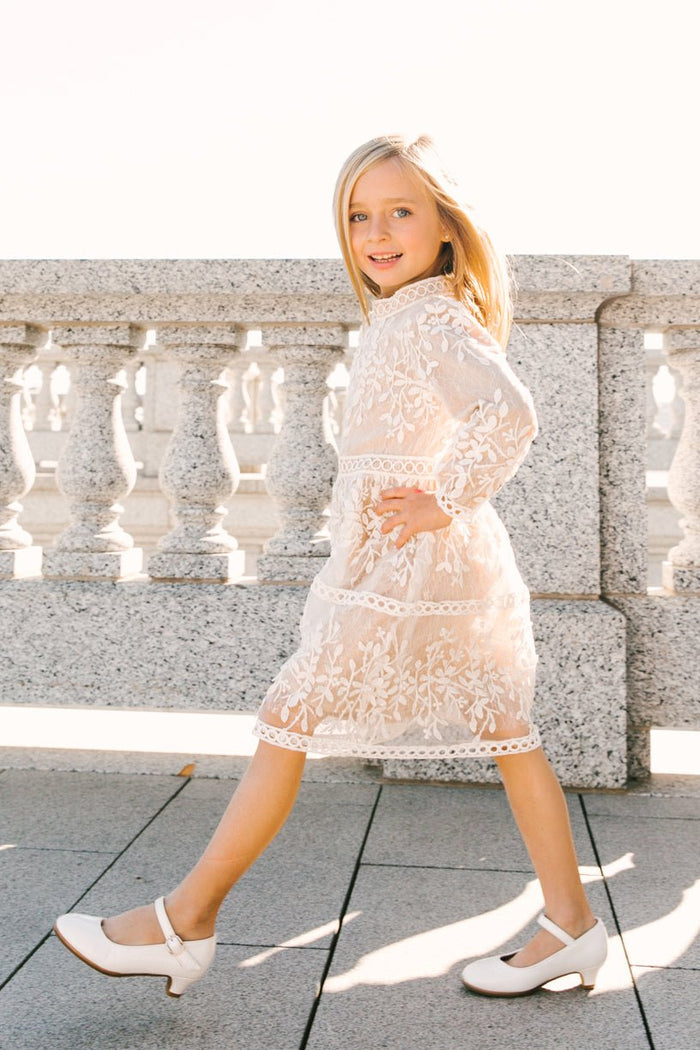 A smiling young girl walks outdoors on a sunny day, wearing the Ivy City Co Mini Sicily Dress with Long Sleeves and white shoes, with a stone railing in the background.