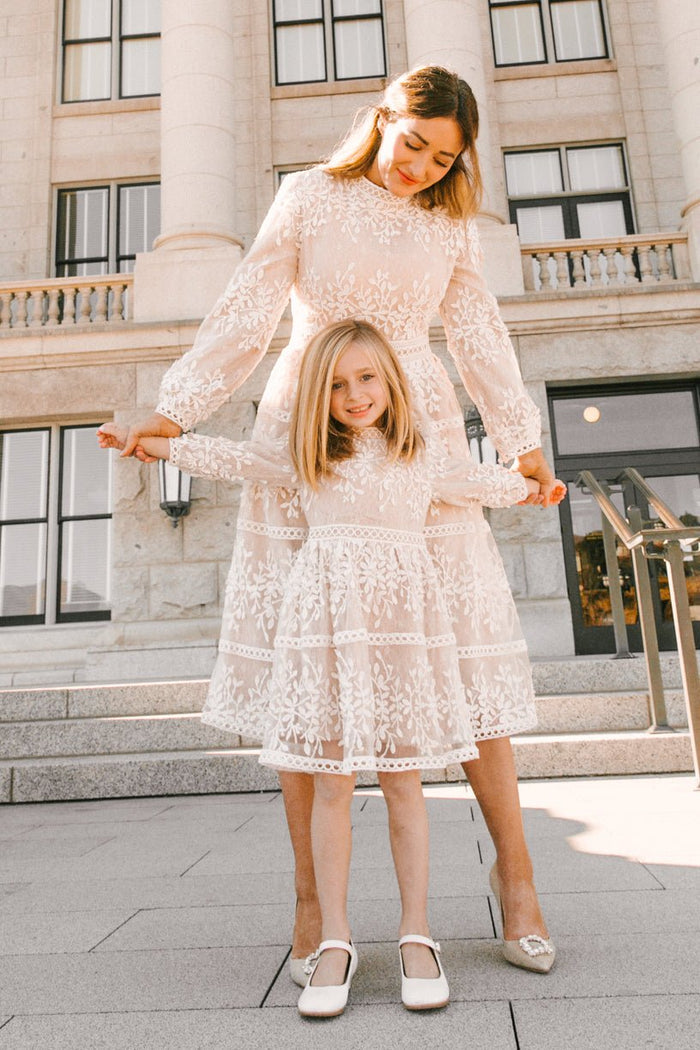 A woman and a young girl, both in elegant white lace dresses by Ivy City Co, stand together holding hands on stone steps in front of a building with columns. The woman wears the brand’s Mini Sicily Dress with Long Sleeves, while the girl’s delicate champagne-lined dress adds a timeless charm to the scene.
