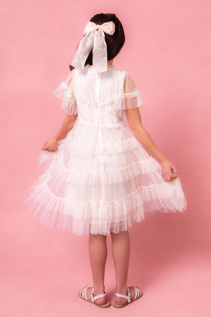 A young girl stands against a pink backdrop, facing away and holding the sides of her Ivy City Co Mini Whimsical Dress in White, featuring flutter sleeves and a layered tulle skirt. She pairs it with white sandals and a large white hair bow.