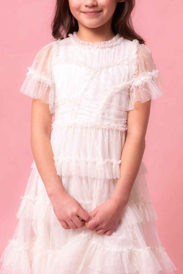 A young girl wears the Ivy City Co Mini Whimsical Dress in White, featuring flutter sleeves and a layered tulle skirt, as she stands against a pink background, smiling softly with her hands clasped. The dress showcases sheer, textured layers and delicate details.