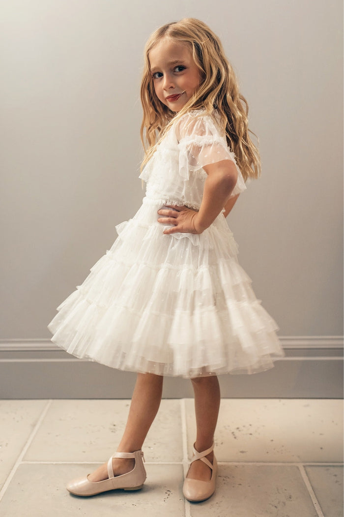 A young girl with long blonde hair poses with one hand on her hip, showcasing the Ivy City Co Mini Whimsical Dress in White, which features a layered tulle skirt. She completes the look with pale pink shoes while standing on a light tile floor against a plain wall.