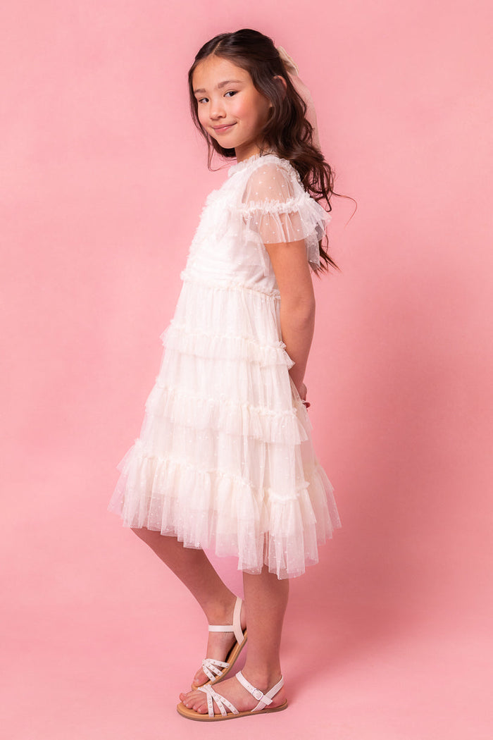 A young girl wearing the Ivy City Co Mini Whimsical Dress in White, showcasing its white ruffled layered tulle skirt, paired with white sandals. She stands sideways against a pink background, her long wavy hair cascading down as she gives a slight smile.