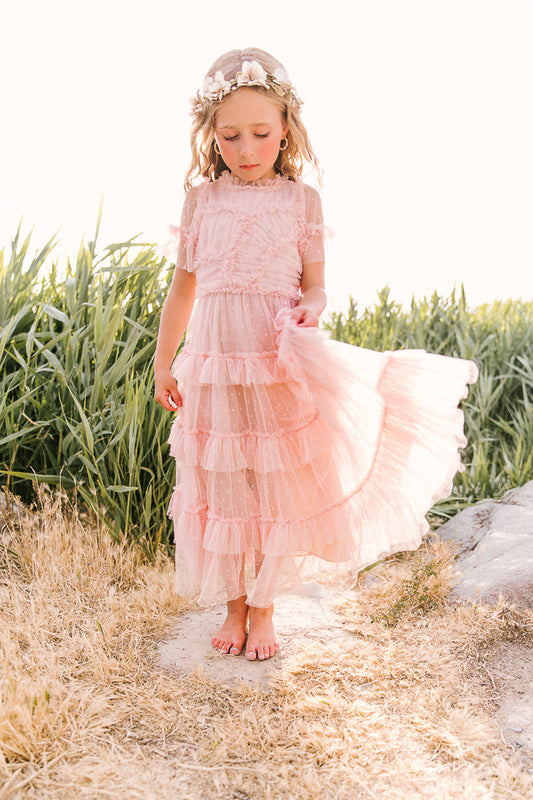 A young girl wears the Ivy City Co Mini Whimsical Maxi Dress in Blush, barefoot on dry grass, lifting one side of her flutter-sleeve tulle skirt. She’s crowned with flowers, surrounded by green plants and bright sunlight.