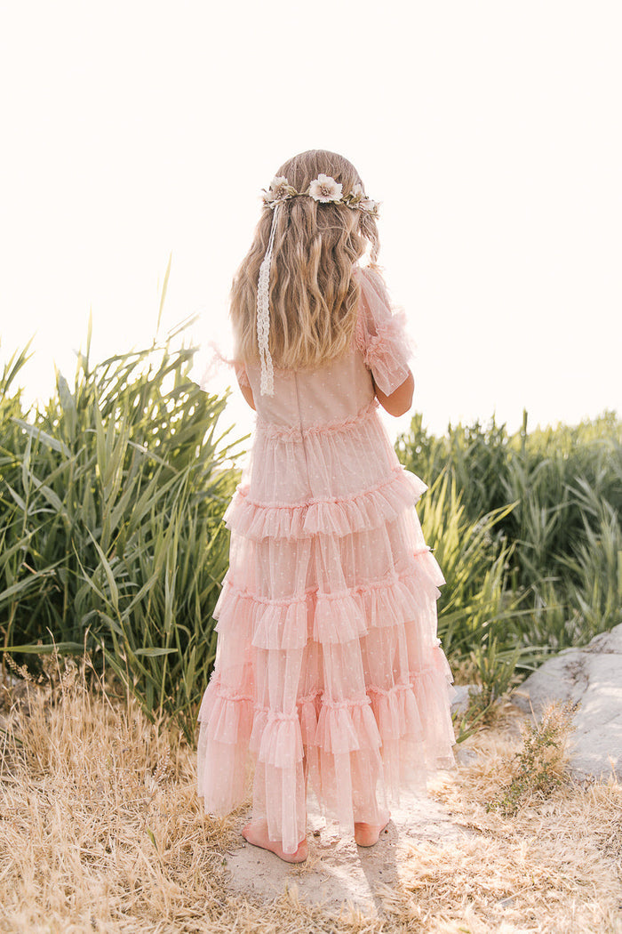 A young girl with long blonde hair, dressed in the Ivy City Co Mini Whimsical Maxi Dress in Blush and a floral headband, stands barefoot on dry grass facing tall green plants, her back to the camera.