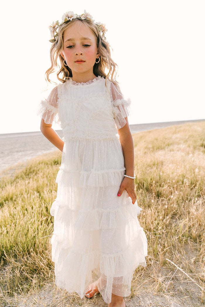 A young girl stands barefoot on dry grass, gazing slightly downward, wearing the Ivy City Co Mini Whimsical Maxi Dress in White with flutter sleeves and a layered tulle skirt. A bright sky and open field provide a dreamy backdrop behind her.