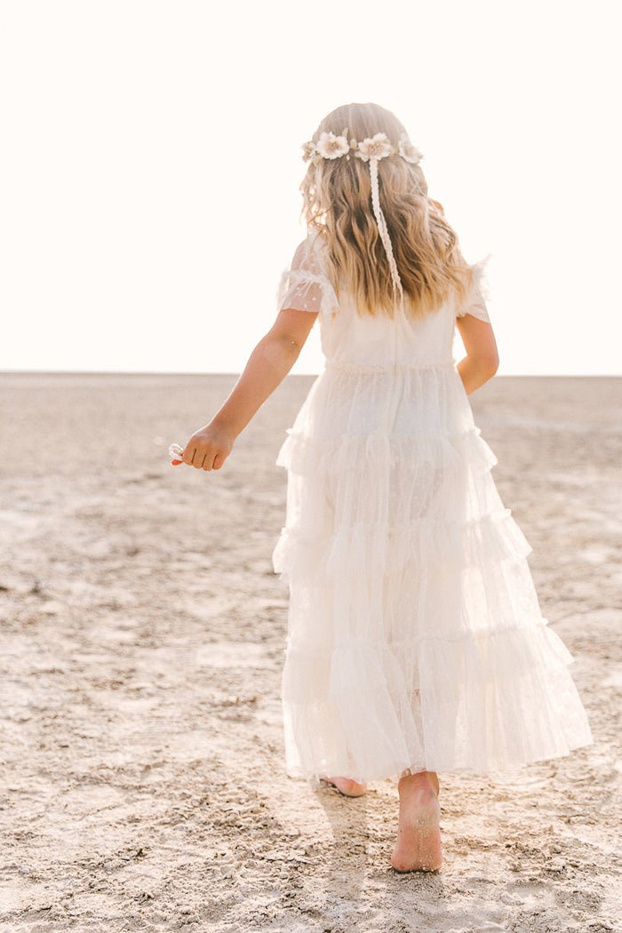 A young girl, barefoot on a sunlit sandy beach and crowned with flowers, faces away from the camera wearing Ivy City Co’s Mini Whimsical Maxi Dress in White—a charming maxi-length piece featuring flutter sleeves and a layered tulle skirt.