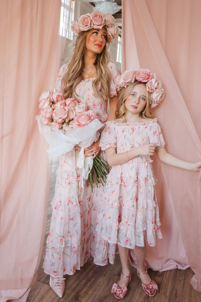 A woman and young girl wear matching Ivy City Co Mini Winnie Dresses in Pink Floral, featuring smocked bodices. Standing before pink curtains with large rose headpieces, the woman holds a bouquet as they exude soft, elegant charm.