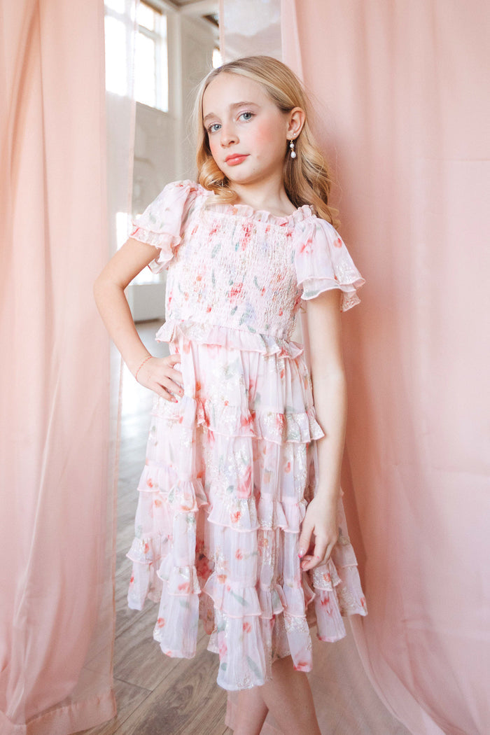 A young blonde girl stands confidently indoors between sheer pink curtains, wearing Ivy City Co’s Mini Winnie Dress in Pink Floral—a smocked-bodice dress—hand on hip and a slight smile.