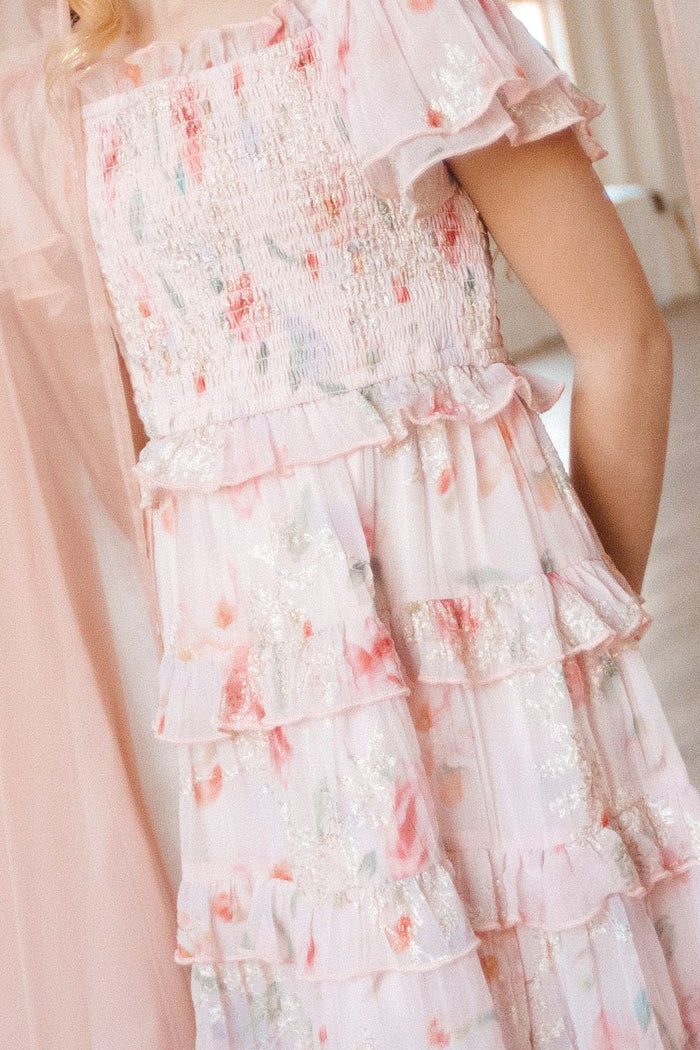 A close-up of a person wearing the Mini Winnie Dress in Pink Floral by Ivy City Co, featuring a smocked bodice, tiered ruffles, glittery accents, and airy fabric for a soft, romantic look.