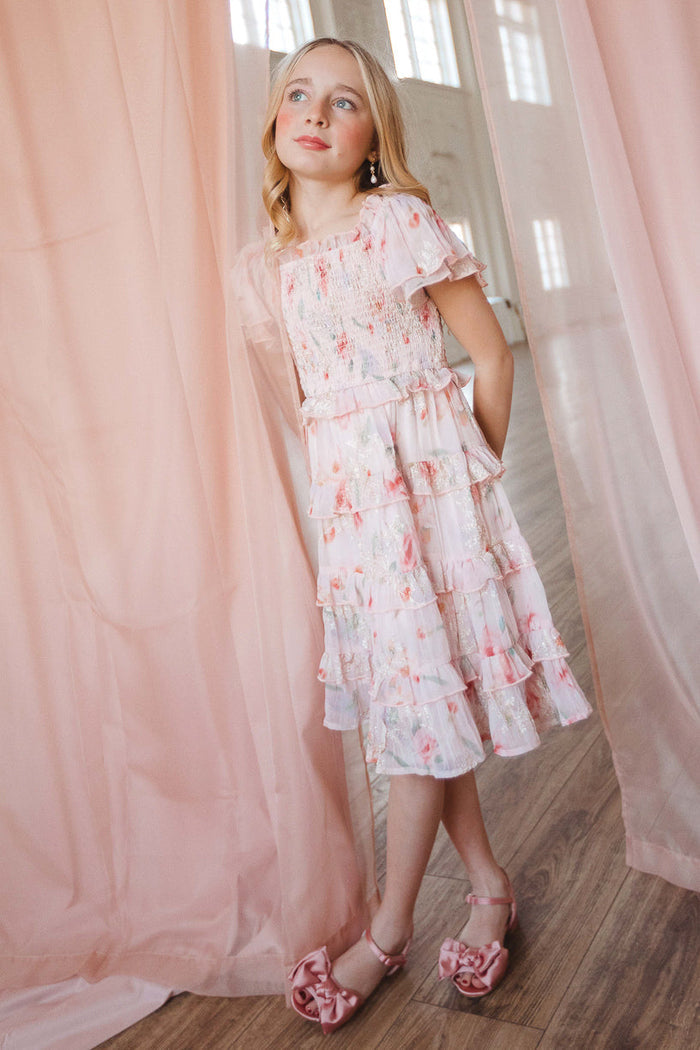 A young girl wearing the Ivy City Co Mini Winnie Dress in Pink Floral and matching sandals stands by sheer pink curtains, gazing upward with a gentle expression in a bright room with wooden floors.