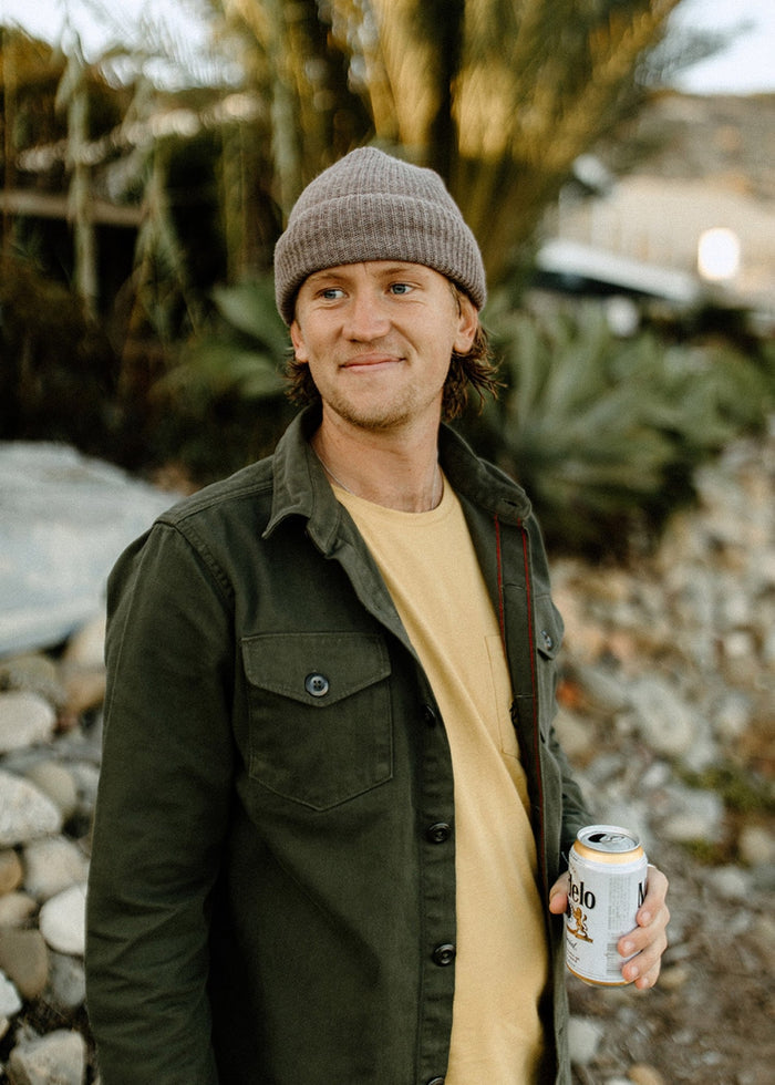 Man in a beanie and green jacket smiles, holding a can outdoors. Palm trees and rocks form the background, creating a relaxed vibe.