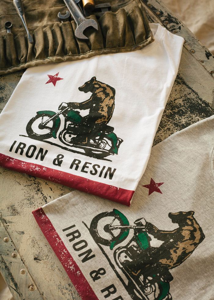 T-shirts with bear on motorcycle design, labeled "Iron & Resin," lie on a rustic table beside a tool roll with wrenches.