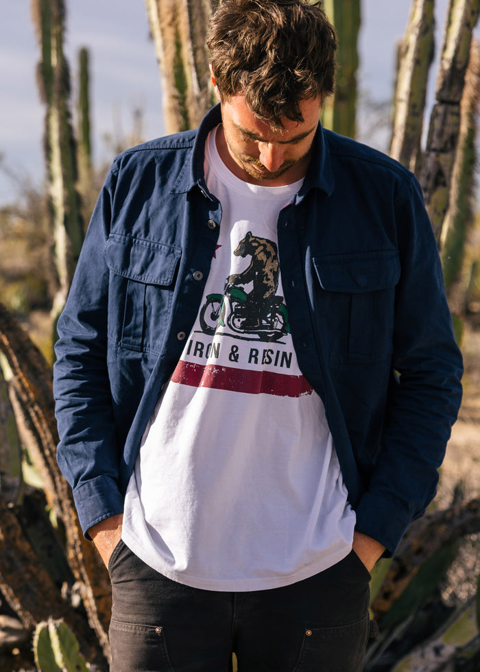 Man in a blue jacket and graphic tee stands in a desert with cacti, looking down. Sunlight highlights the shirt's design.