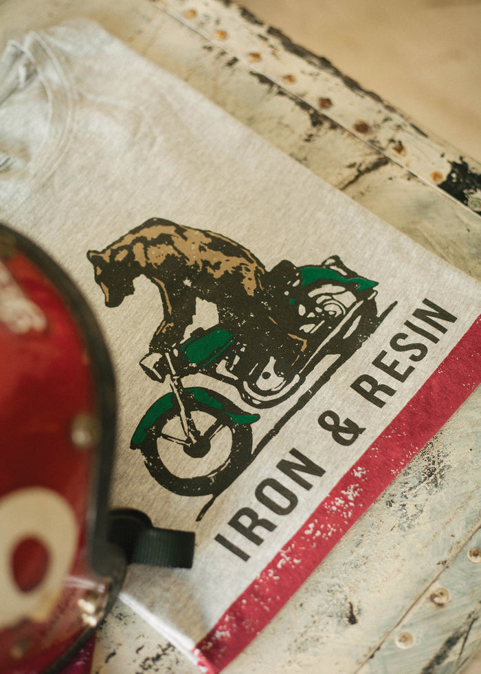 T-shirt with a bear on a motorcycle design, labeled "IRON & RESIN," lies on a rustic table beside a red helmet.