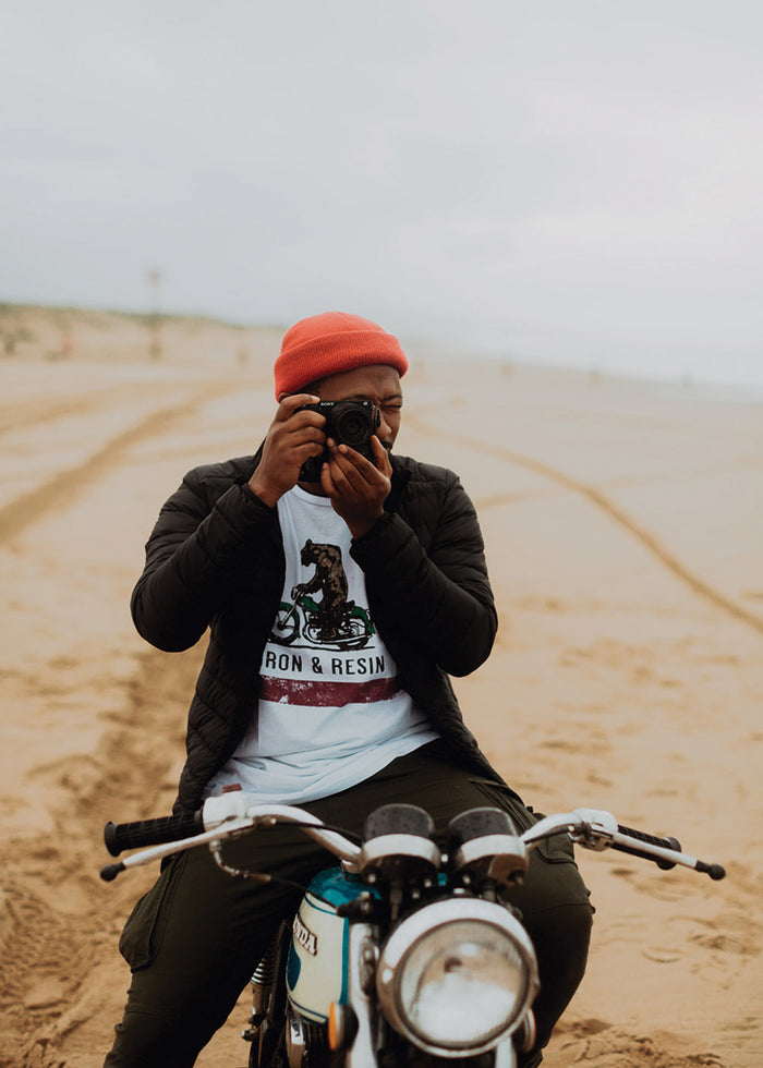 Man in red beanie takes photo on beach, sitting on vintage motorcycle, wearing a graphic tee and black jacket.