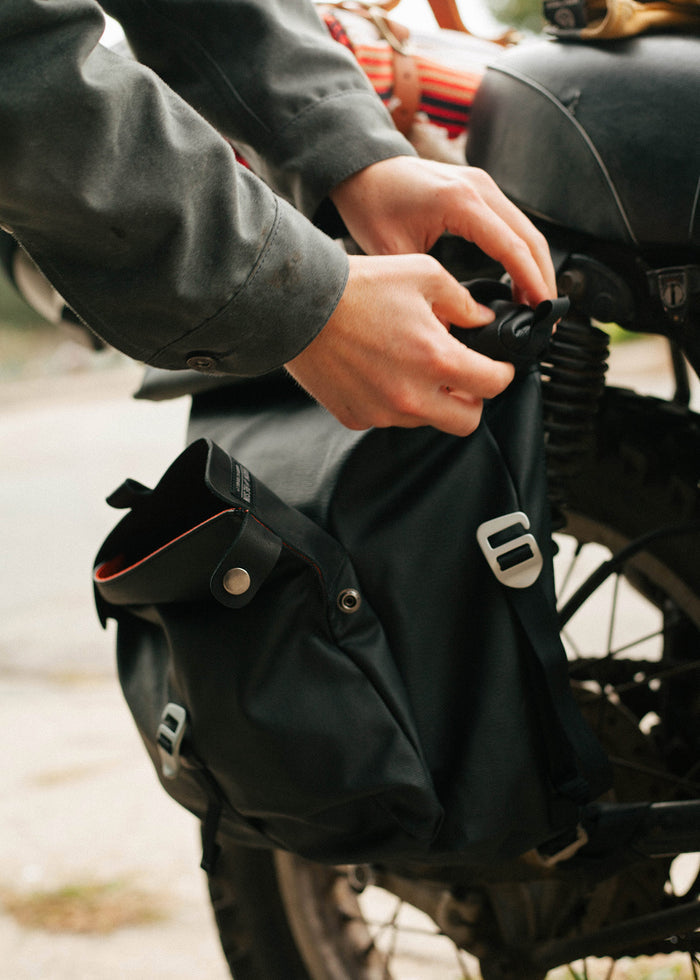 Hands adjusting a black saddlebag on a motorcycle, showcasing travel gear and adventure preparation.