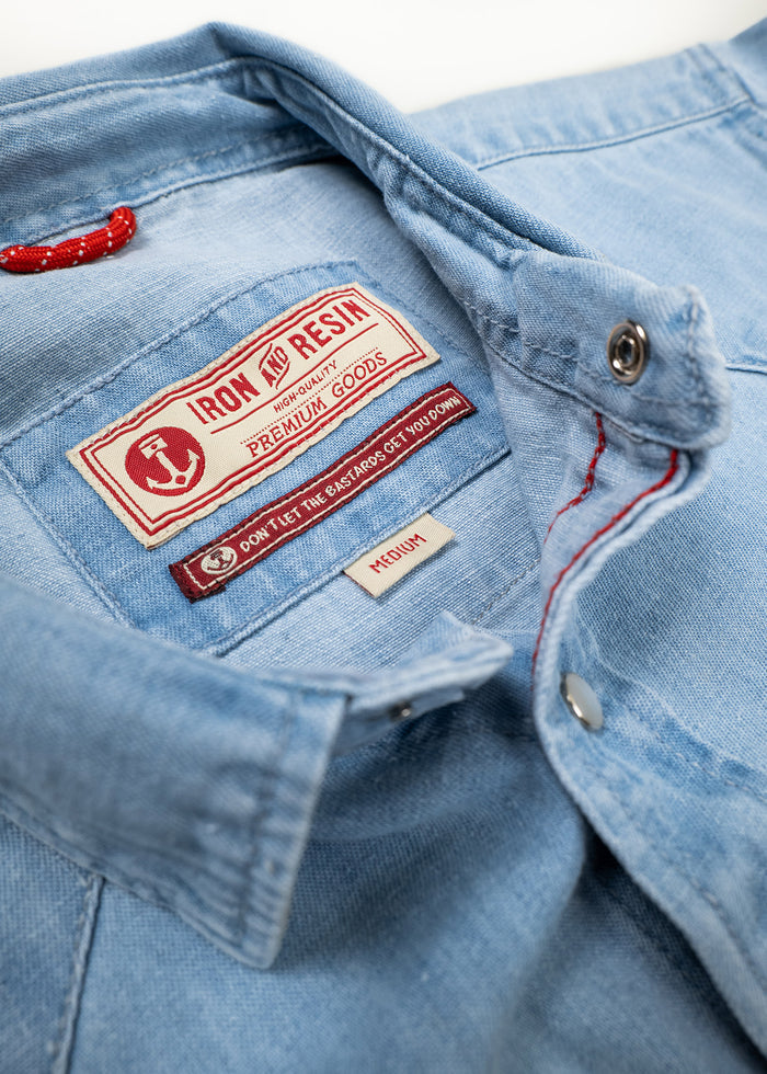 Close-up of a light blue denim shirt with "Iron and Resin" label, highlighting premium quality and medium size.