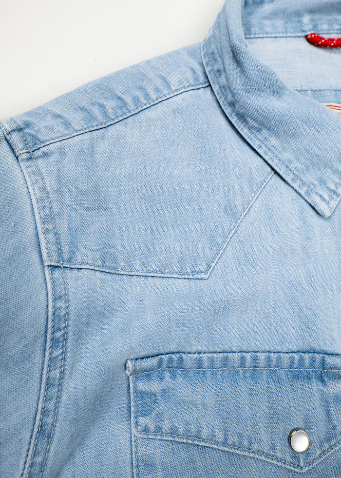 Close-up of a light blue denim shirt with a pointed collar and a buttoned chest pocket, showcasing its texture and stitching details.