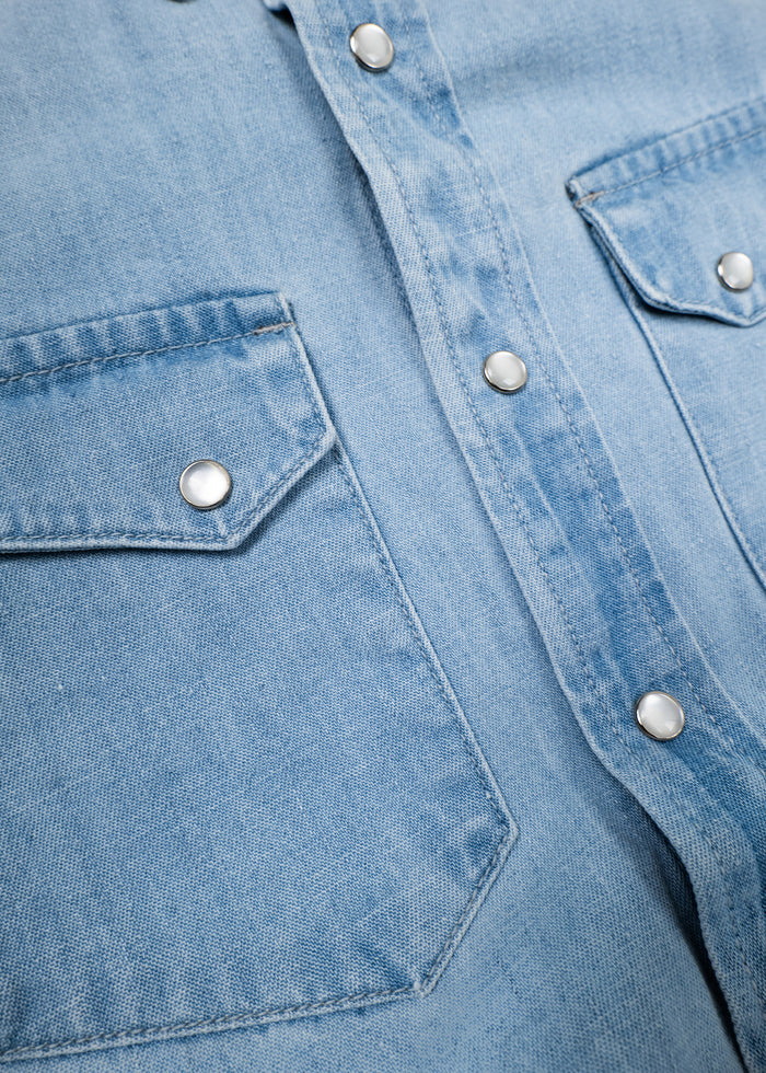 Close-up of a light blue denim shirt with silver snap buttons and two chest pockets, highlighting texture and stitching details.