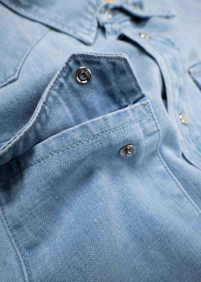 Close-up of a light blue denim shirt with metal snap buttons and a visible pocket, highlighting texture and stitching details.