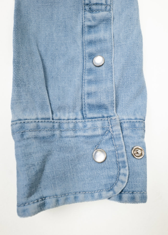 Close-up of a light blue denim sleeve with two silver snap buttons on a white background, highlighting casual fashion details.