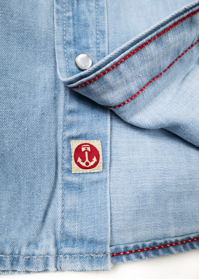 Light blue denim shirt with red stitching and a silver button. A small red anchor logo patch is visible near the hem.