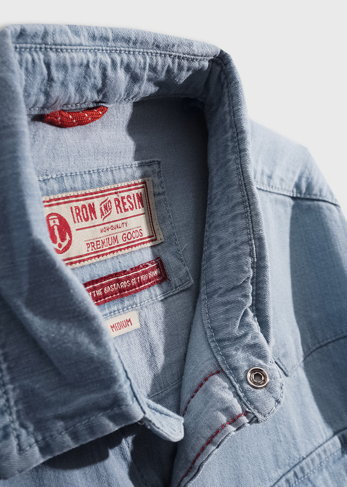 Close-up of a light blue denim shirt collar with red stitching, featuring an Iron and Resin label indicating premium quality goods.