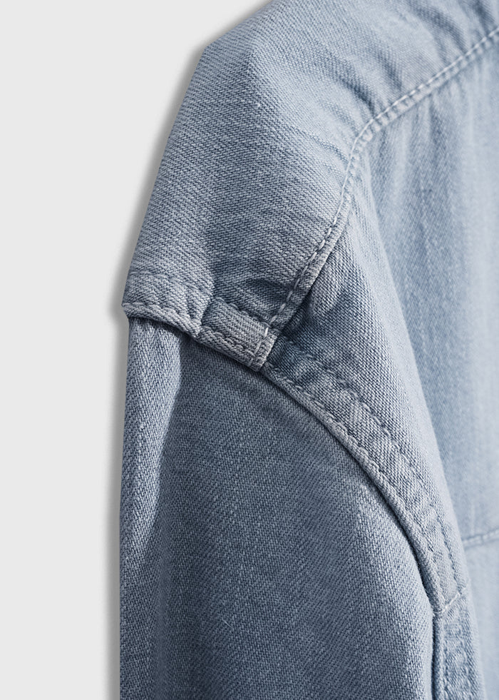 Close-up of a blue denim jacket's shoulder seam, highlighting the texture and stitching detail on a light background.