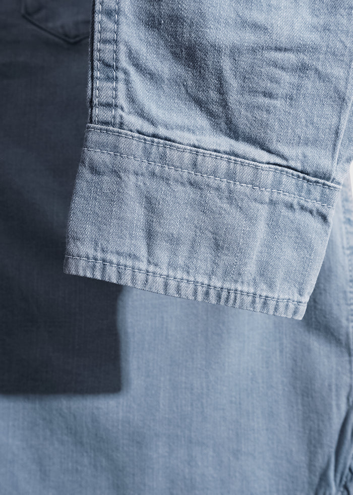 Close-up of a light blue denim shirt sleeve, showing detailed stitching and texture under natural light.