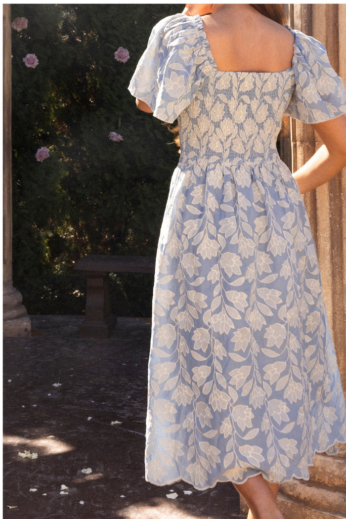 A woman stands outdoors in a sunlit garden, her back turned, wearing the Ivy City Co Ophelia Dress in Mosaic Embroidery. The dress features puffed sleeves and a smocked waist, with stone columns and lush greenery behind her.