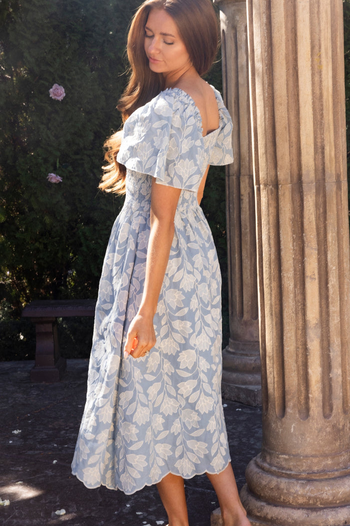 A woman in the Ivy City Co Ophelia Dress in Mosaic Embroidery stands barefoot by a stone column, looking down, with greenery and a bench in the background.