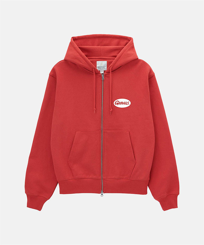Model wearing a Gramicci crimson red Oval Patch Zip Hooded Sweatshirt with a red and white patch against a light-tone background.