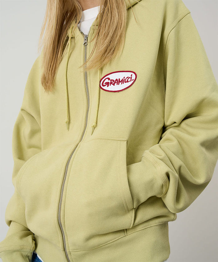 Model wearing a Gramicci lime Oval Patch Zip Hooded Sweatshirt with a red and white patch  against a light-tone background.