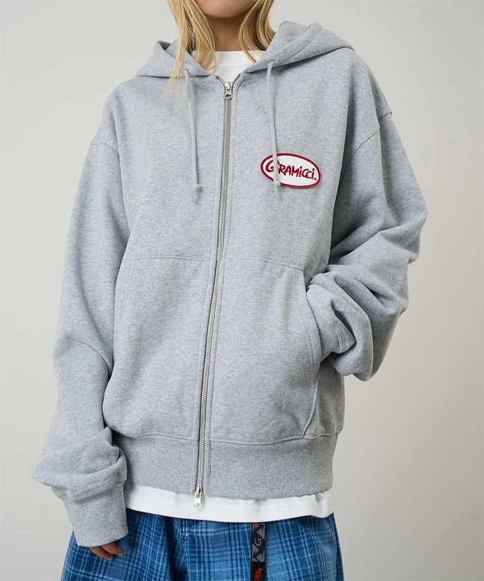 Model wearing a Gramicci heather grey Oval Patch Zip Hooded Sweatshirt with a red and white patch against a light-tone background.