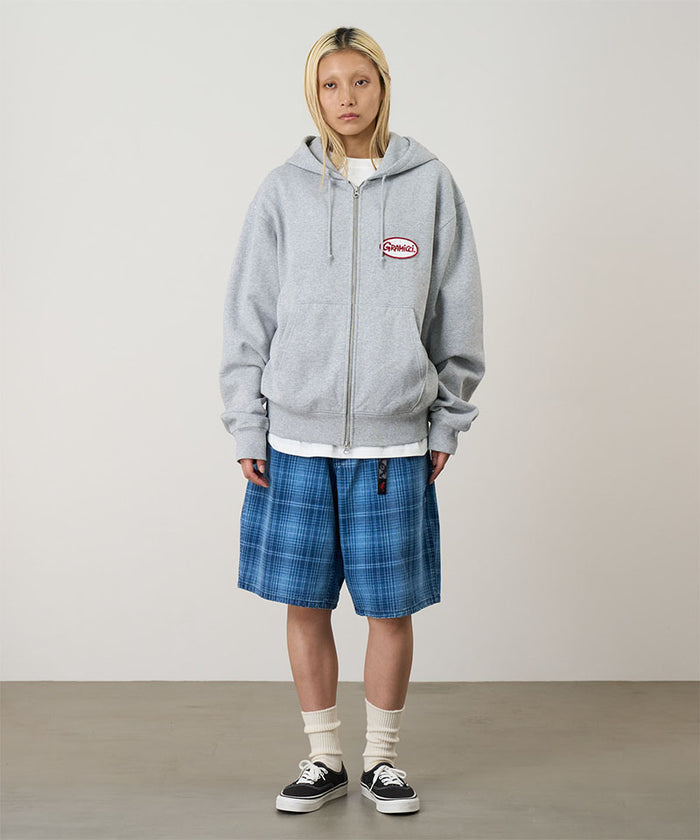 Model wearing a heather grey Gramicci Oval Patch Zip Hooded Sweatshirt with a red and white patch and blue plaid shorts on a light-tone background.