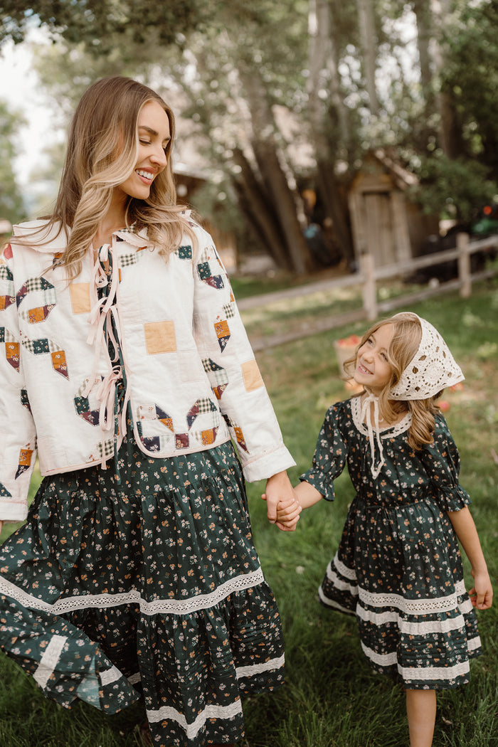 A woman and young girl wearing matching Ivy City Co Palermo 3/4 Sleeves Dresses in Evergreen Lovelorn Ditsy hold hands and smile at each other outdoors. The woman’s dress offers a comfortable 3/4 sleeve design with a maternity-friendly fit, making it perfect for any stage. The girl accessorizes with a crocheted bonnet, while trees and a wooden fence appear in the background.